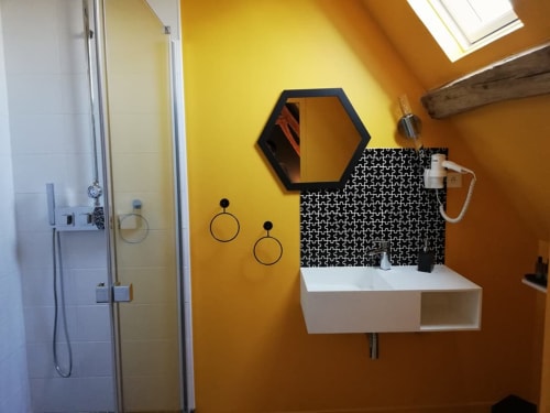 Salle de bain moderne à La Cour d'Hortense : murs jaunes, miroir hexagonal, lavabo blanc, dosseret à motifs noirs et verre