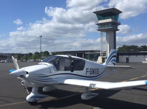 Petit avion blanc aux bandes bleues garé à l'aérodrome de Sailly-Flibeaucourt avec tour de contrôle visible