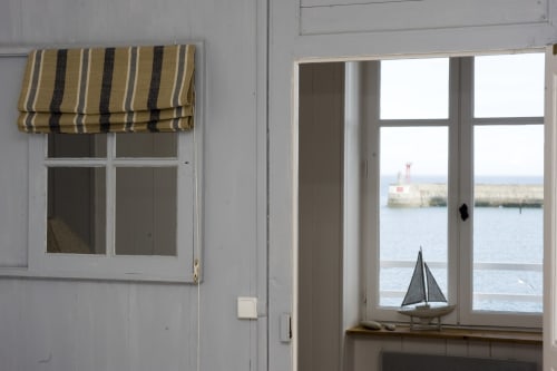 Chambre de maison côtière avec store rayé, fenêtre blanche donnant sur le port de Port-en-Bessin, phare et voilier