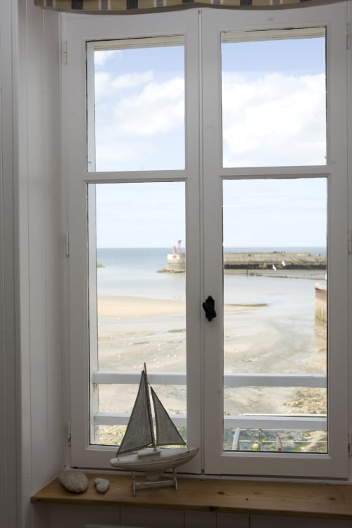 Fenêtre de chaumière côtière donnant sur le port de Port-en-Bessin avec voilier et vue sur la plage de sable