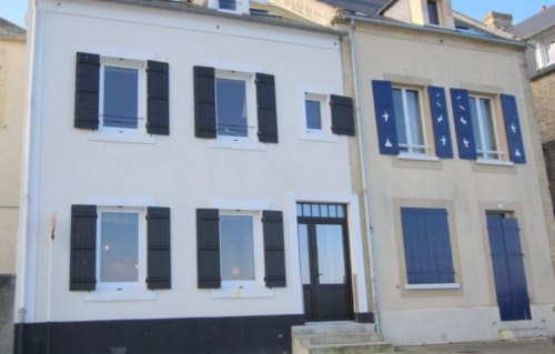 Façade de cottage blanc et beige aux Filles du Bord de Mer à Port-en-Bessin avec volets noirs et portes bleues