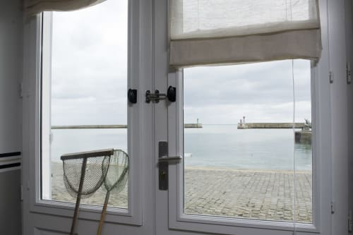Open glass doors view of Port-en-Bessin harbor with fishing nets and stone pier, Les Filles du Bord de Mer
