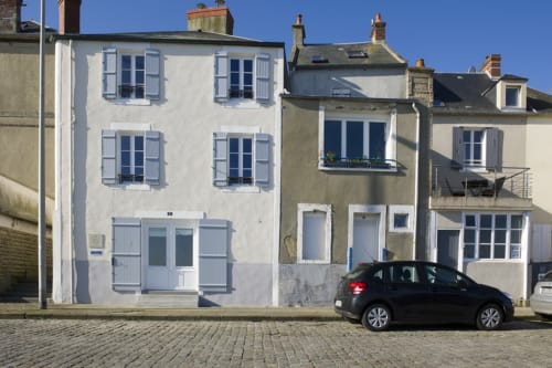 Witgekalkt huisje aan Les Filles du Bord de Mer in Port-en-Bessin met blauwe luiken en geparkeerde auto