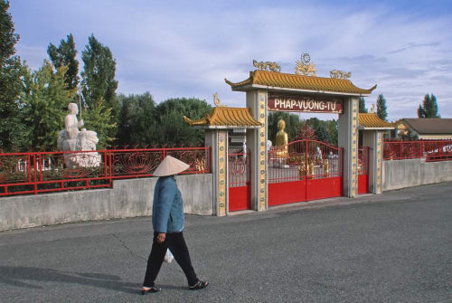 Person in blue jacket and conical hat walking toward ornate red and gold Asian-style temple gate at Château De Clusors in