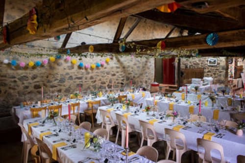 Salle de banquet en pierre rustique à La Raymondière avec nappes blanches, touches jaunes et décors en papier coloré suspendus.