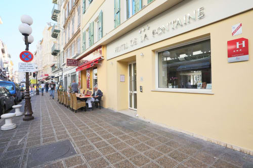 Hôtel De La Fontaine entrance on a sunny Nice street with outdoor seating and pedestrians