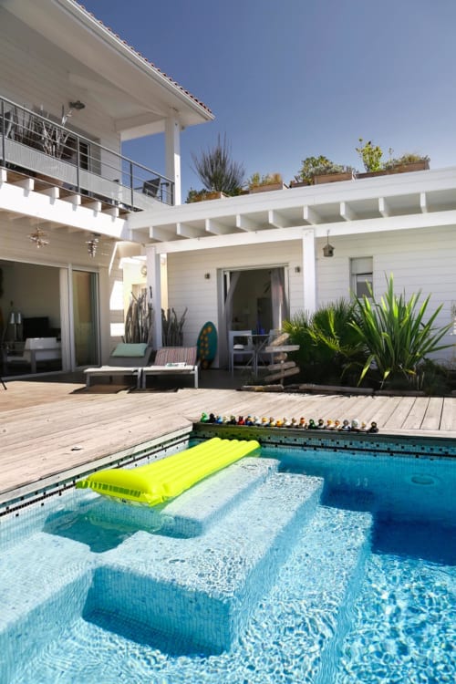 Vue lumineuse de la piscine de l'hôtel Côte Et Dune à Biscarrosse Plage avec bâtiment blanc moderne, terrasse en bois et piscine bleue