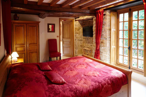 Chambre luxueuse du Moulin d'Ajon avec parure velours rouge, murs en pierre apparente, poutres bois et portes-fenêtres