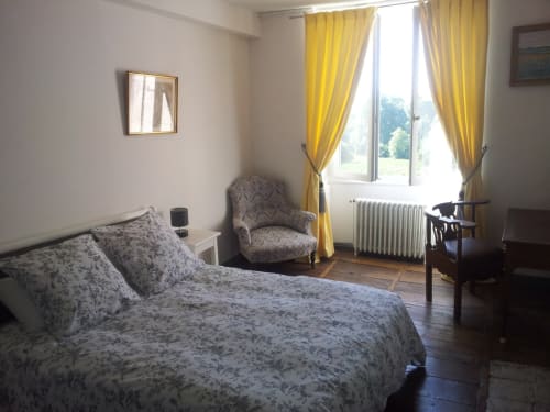 Chambre Aymard au Domaine De Puyrousse avec literie fleurie, fauteuil, rideaux jaunes et vue sur le jardin