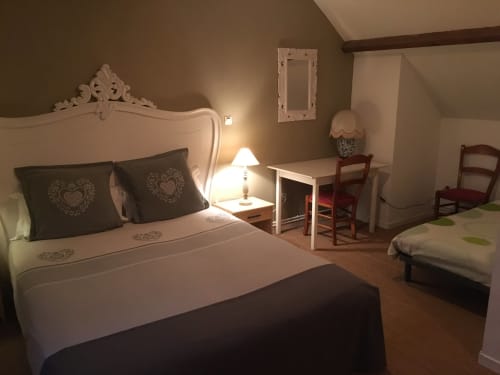 Chambre élégante à La Ferme Du Grand Air avec lit blanc orné, oreillers sombres, lampe de chevet et mobilier en bois à Fiennes