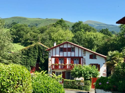 Hotel Au Bon Coin in Biarritz, a red and white Basque-style building nestled in lush green hills with mountains in the