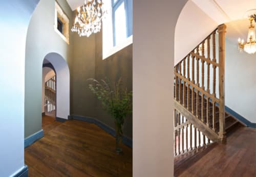 Élégant couloir du Château de Baylac avec escalier en bois, portail voûté, lustre et murs aux bordures bleues.