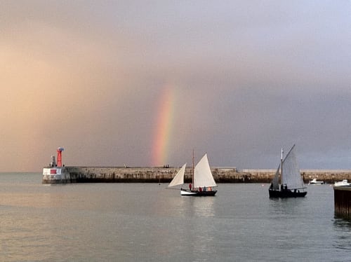 Bateaux à voiles traditionnels au port de Port-en-Bessin avec arc-en-ciel et brise-lames, scène côtière normande