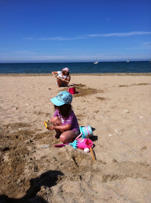 Des enfants construisent des châteaux de sable sur la plage des Filles du Bord de Mer à Port-en-Bessin, avec des voiliers au large
