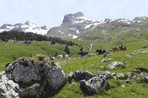 Prairie alpine avec affleurements rocheux, montagnes enneigées, cavaliers et sapins près de Bugnein