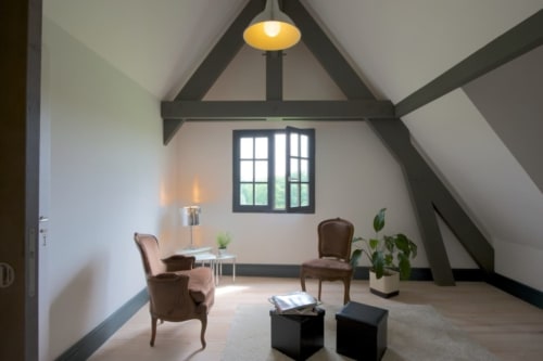 Suite Vivaldi au Château de Baylac : Chambre mansardée contemporaine avec poutres apparentes, fauteuils marron et vue sur les montagnes par baie cintrée