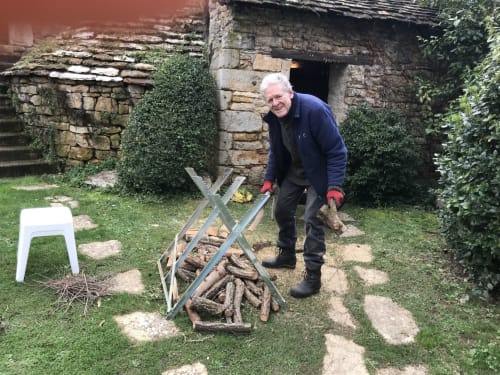 Elderly man with white hair cutting firewood at Manoir De Rieuzal in Loubressac, France, with stone buildings and ivy in