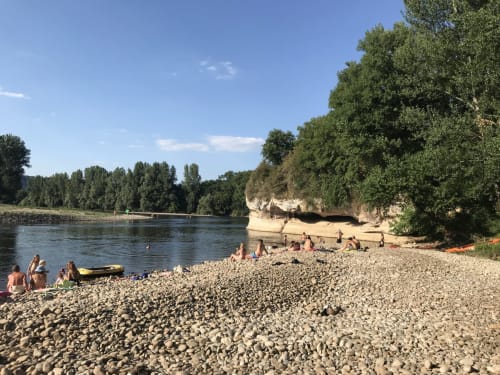 Pebble beach on river with swimmers and boats, limestone cliffs, trees, near Loubressac