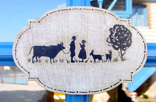 Wooden sign at Au Petit Tambour D'Autingues featuring pastoral silhouettes of farmers, livestock, and a tree in the garden