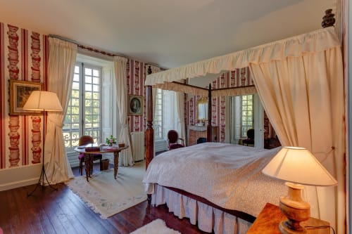 Chambre à baldaquin élégante au Château d'Alexis de Tocqueville avec rideaux crème, papier peint rayé rouge et mobilier d'époque.