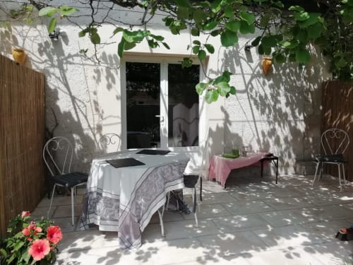 Cour ombragée du Mas des Artistes avec table de repas, chaises, porte blanche, plantes grimpantes et fleurs roses