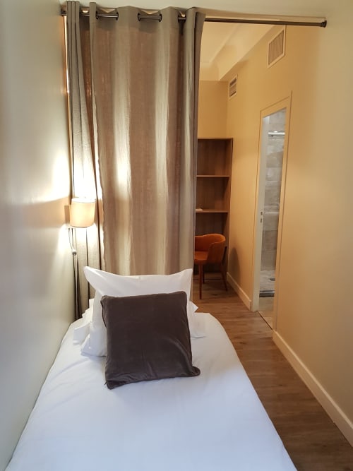 Chambre triple moderne à l'Hôtel Au Bon Coin à Biarritz avec lit blanc, oreillers marron, rideaux beige, parquet et vue sur la ville