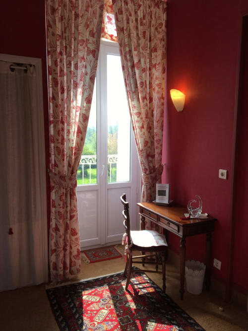 La Chatellenie standard room with red walls, floral curtains, garden view doors, desk, and period furniture