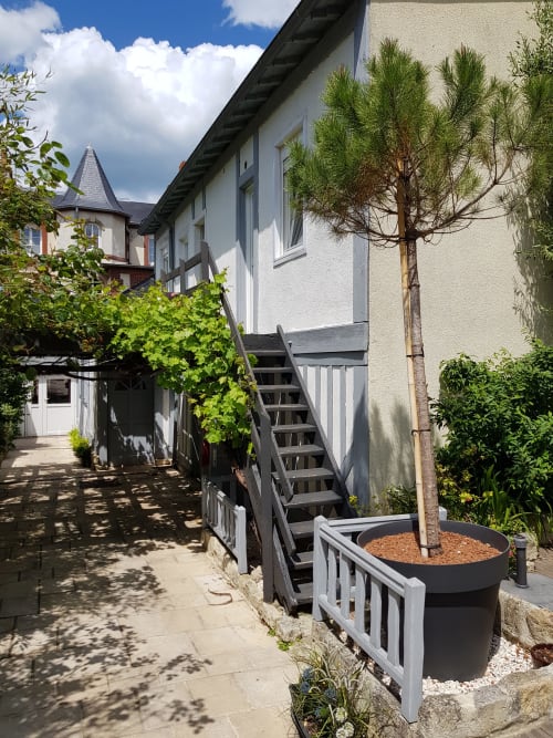 Extérieur de l'hôtel Les Cabines à Houlgate avec escalier en bois, bâtiment aux finitions blanches, sapin en pot et vues sur le jardin luxuriant