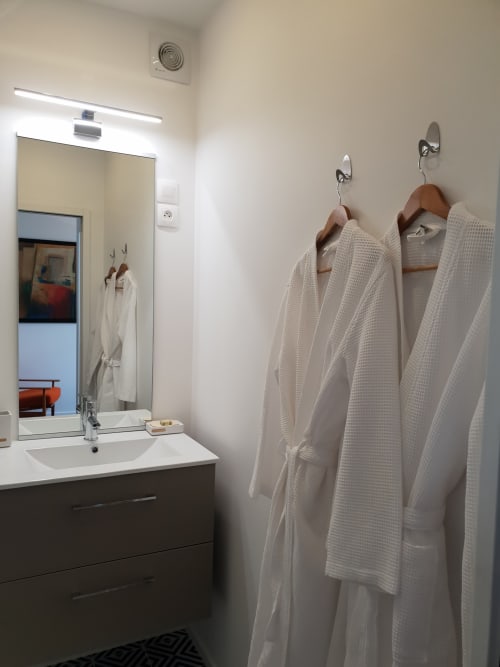 Salle de bain moderne avec lavabo, miroir, éclairage et trois peignoirs blancs à nid d'abeille suspendus aux crochets à La Conciergerie de Nathalie