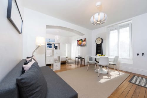 Studio moderne à Un Air De Famille B&B à Vittel avec canapé gris, étagères blanches, coin repas et grandes fenêtres lumineuses