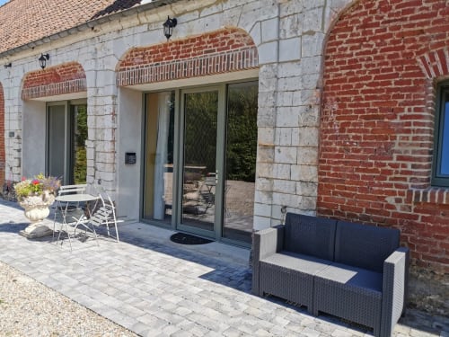 Entrée du gîte La Cour d'Hortense avec murs de brique, portes vitrées, salon de jardin et vue sur le jardin à Sailly-Flibeaucourt