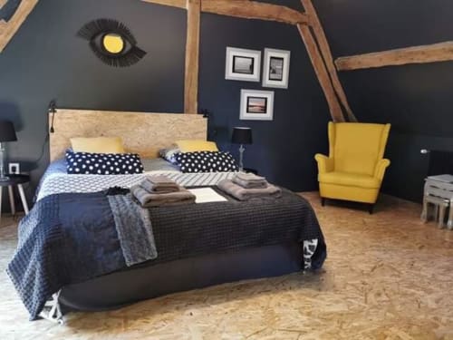 Chambre moderne à La Cour d'Hortense avec murs foncés, poutres en bois, fauteuil jaune et lit aux coussins à motifs