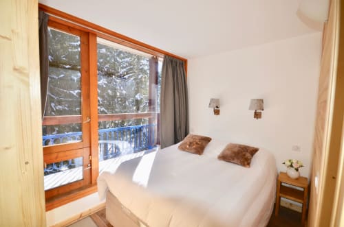 Chambre lumineuse dans un appartement de ski à ARC 1800 avec lit double, vue sur les sapins depuis la porte-fenêtre en bois et forêt enneigée
