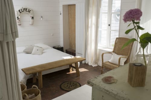 Helles Küstenschlafzimmer mit weißer Schiffsbeplankung, Holzbett, Korbsessel und violetter Blume in Port-en-Bessin