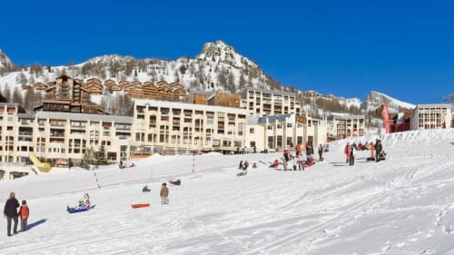 Pente enneigée avec familles en luge, bâtiments alpins et montagnes enneigées dans une station de ski