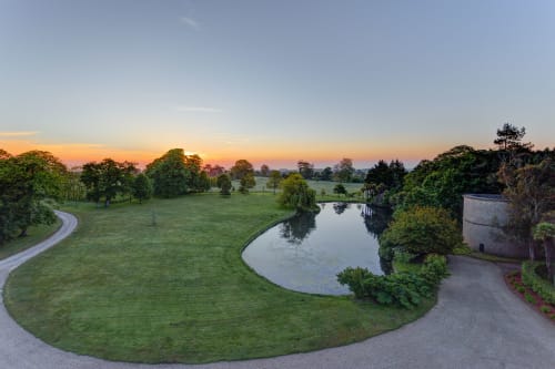Sunrise over Château d'Alexis de Tocqueville grounds with curved pathway, manicured lawns, and reflective pond in Normandy.