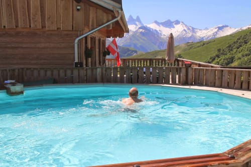 Invité se baignant dans la piscine turquoise du Chalet La Louvière avec les Alpes enneigées et le drapeau suisse en arrière-plan