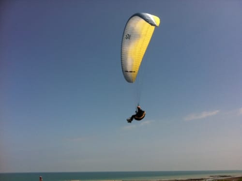 Parapentiste au parachute jaune et blanc survolant la plage de Port-en-Bessin-Huppain et ses eaux turquoise