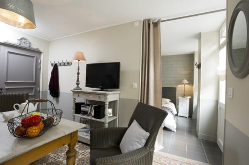 Coin salon cosy de l'appartement Les Filles du Bord de Mer avec téléviseur, fauteuil en osier, panier de fruits et vue sur la chambre