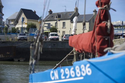 Bateau de pêche bleu avec bouée rouge au port de Port-en-Bessin, bâtiments en pierre et voitures garées en arrière-plan