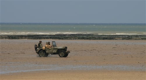 Jeep militaire sur la plage de Port-en-Bessin avec jetée historique et vagues en arrière-plan