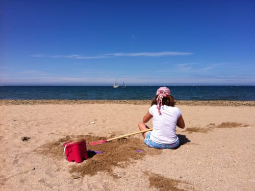 Enfant jouant au seau et à la pelle sur la plage de sable de Port-en-Bessin avec des voiliers au large