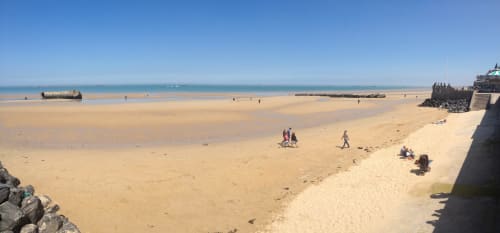 Plage de sable à Port-en-Bessin avec visiteurs, digues et bateaux à l'horizon