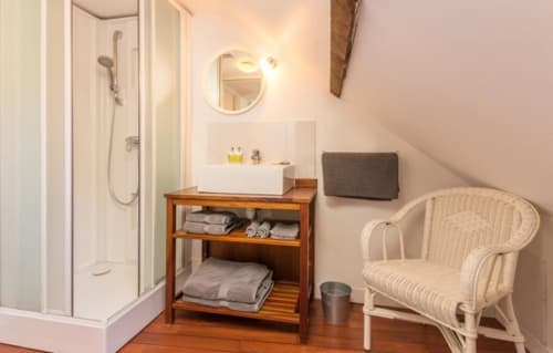 Salle de bain lumineuse du gîte Les Filles du Bord de Mer avec douche, meuble vasque en bois, miroir, fauteuil en osier et charme côtier
