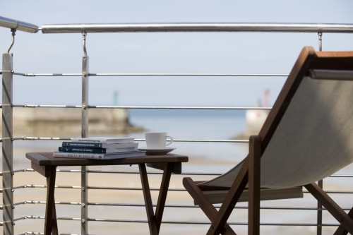 Terrasse en bois avec vue sur mer, livres, tasse de café et balustrade moderne donnant sur la plage de Port-en-Bessin