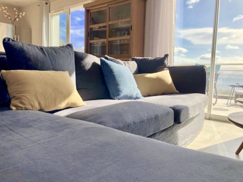 Coastal villa lounge with sea-view windows, gray upholstered sofa, blue and beige accent pillows, wooden cabinet, and sunny