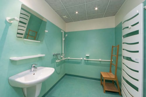 Salle de bains accessible turquoise vif à Un Air De Famille avec barres d'appui, douchette et lavabo blanc à Vittel.