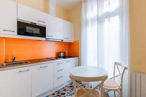 Modern white kitchen with vibrant orange backtile, dining table, and natural light at Hotel Residence Europe & Spa in Clichy