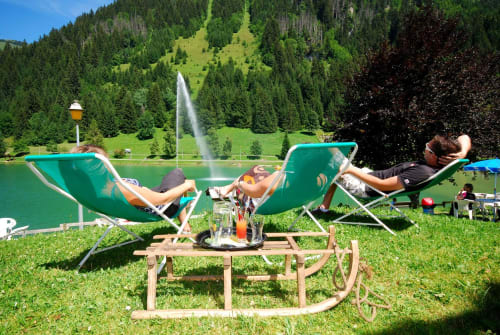 Détente au bord du Lac de Vonnes, jet d'eau, été, chaises longues, forêt de sapins, paysage verdoyant. (Page Châtel-Portes-du-Soleil)