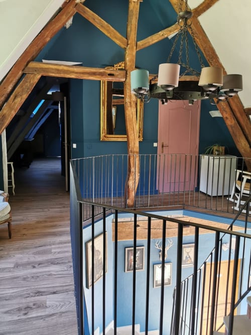 Chambre rustique en mezzanine à La Cour d'Hortense avec poutres en bois apparentes, murs bleu-vert et garde-corps métalliques.